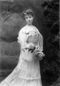 A black and white photograph of Elizabeth von Arnim, wearing a white dress and holding a bouquet of flowers